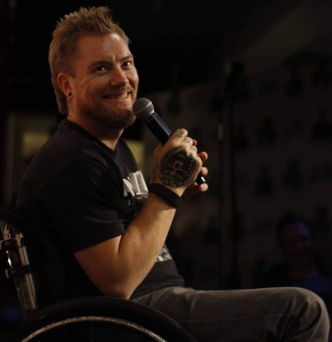 Kevin Brooks on stage in his wheelchair at a comedy show, microphone in one tattooed hand, grinning at the audience mid-set