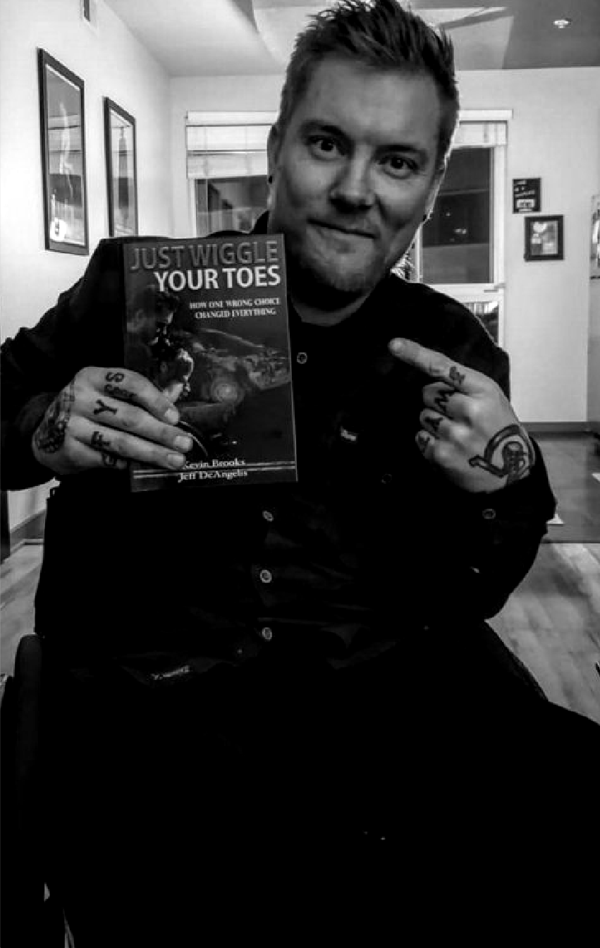 Kevin Brooks at home holding up a copy of his book, Just Wiggle Your Toes, and pointing at the cover with a tattooed hand
