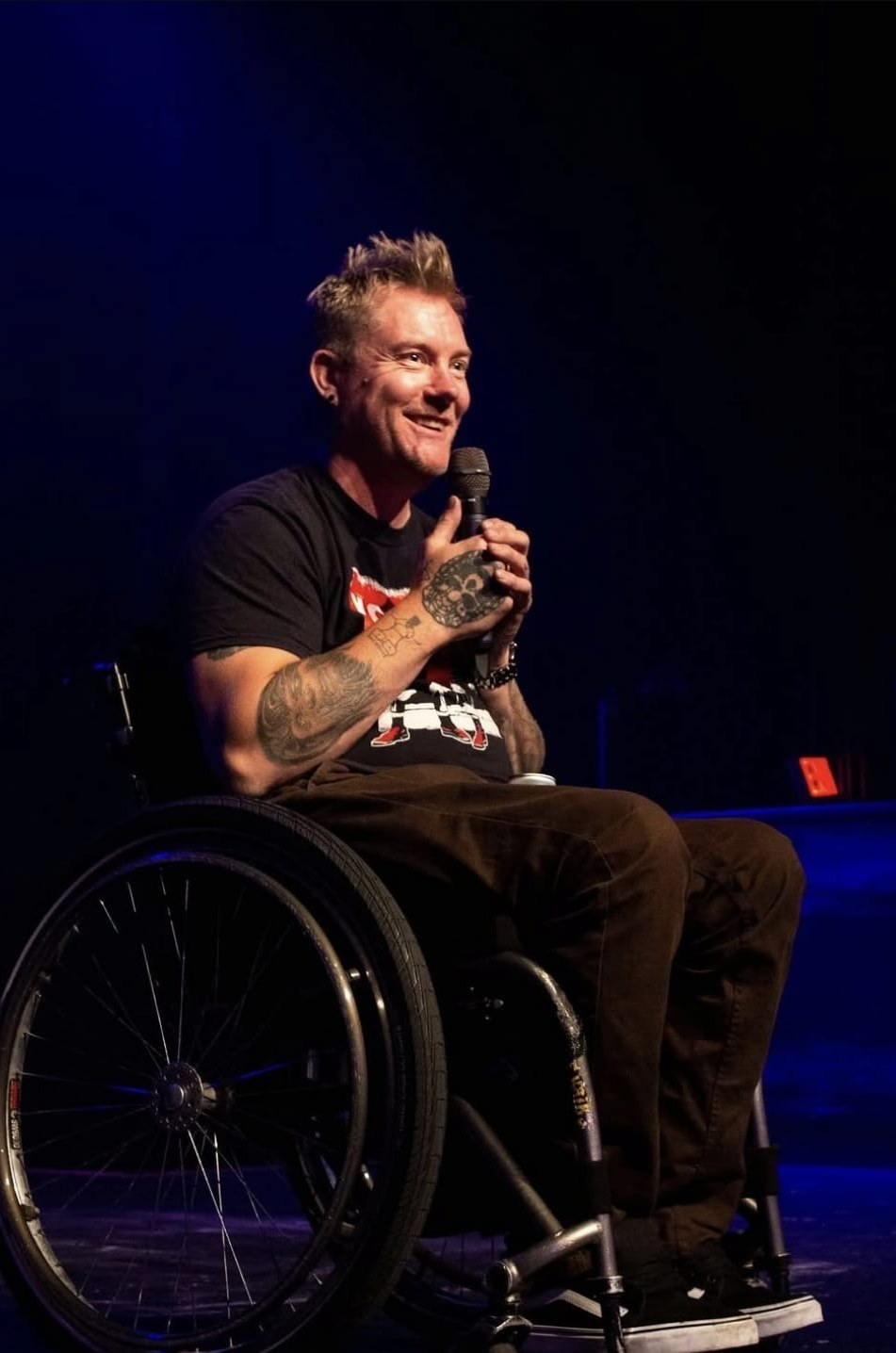 Kevin Brooks on stage in his wheelchair, microphone in one tattooed hand, grinning out at the room under a blue stage wash