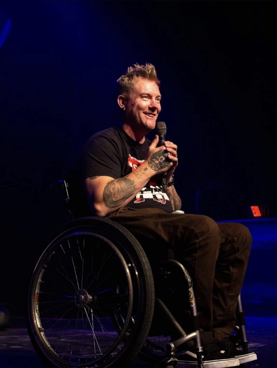 Kevin Brooks on stage in his wheelchair at the Rickshaw in Vancouver, microphone in one tattooed hand, grinning out at the room under a blue stage wash