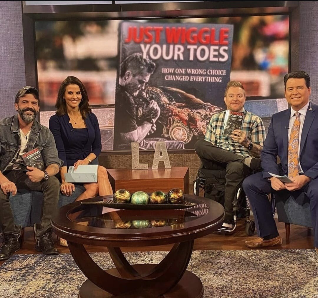 Kevin on a daytime television set holding his book, beside a host, a co-host and another guest, the full cover of Just Wiggle Your Toes filling the screen behind them